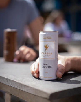 Hands holding Cafeda Vietnamese iced coffee cans alongside fresh baguette sandwiches, showcasing pre