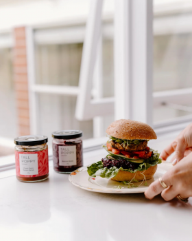 Hand holding Tall Poppy Gourmet Classic Spicy Tomato Relish jar beside sliced tomatoes and cucumber