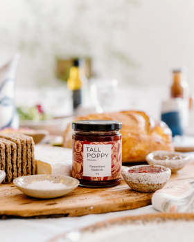 Hand holding Tall Poppy Gourmet Classic Spicy Tomato Relish jar beside sliced tomatoes and cucumber